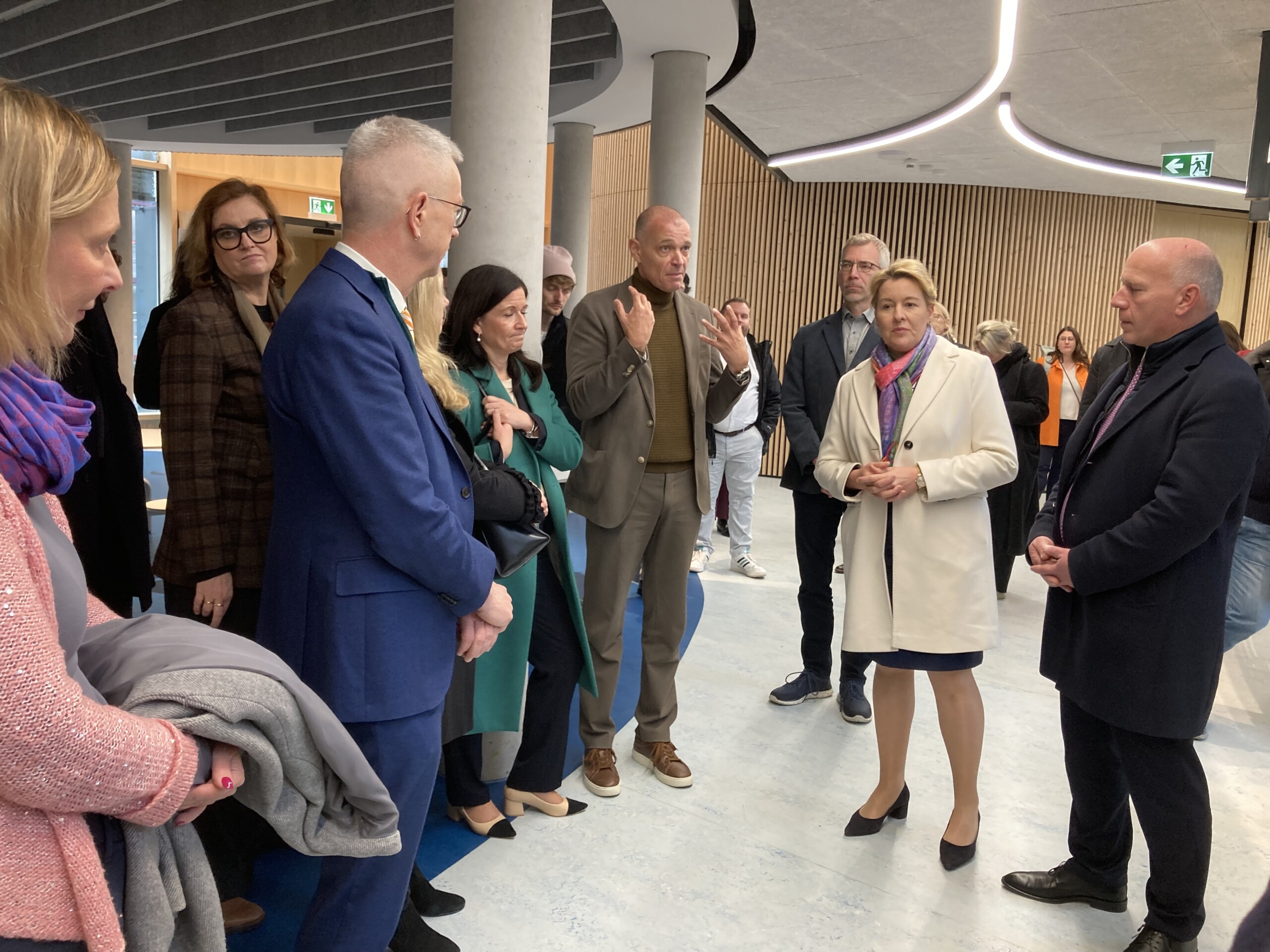 Gruppe von Personen in einem modernen Innenraum, darunter Roland Sillman im braunen Anzug, Franziska Giffey in weißem Mantel und Kai Wegner in dunklem Anzug, stehend in Gespräch.